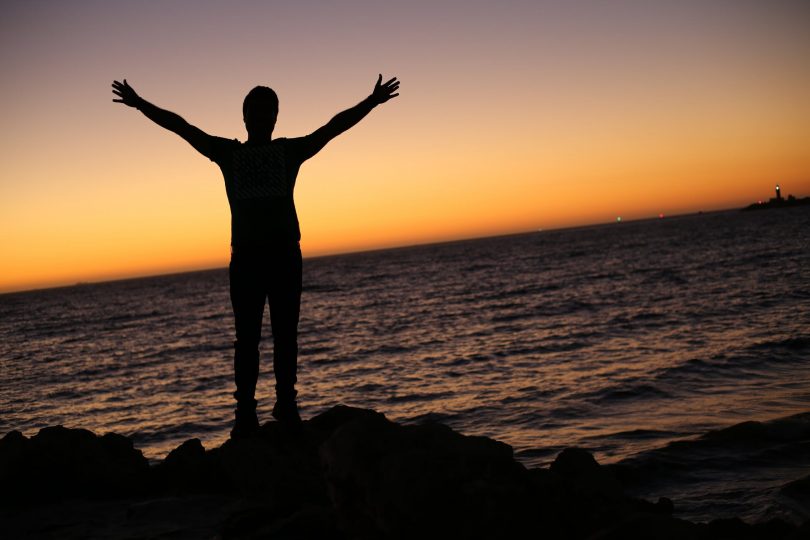 Silhouette of person standing with arms raised in front of ocean.