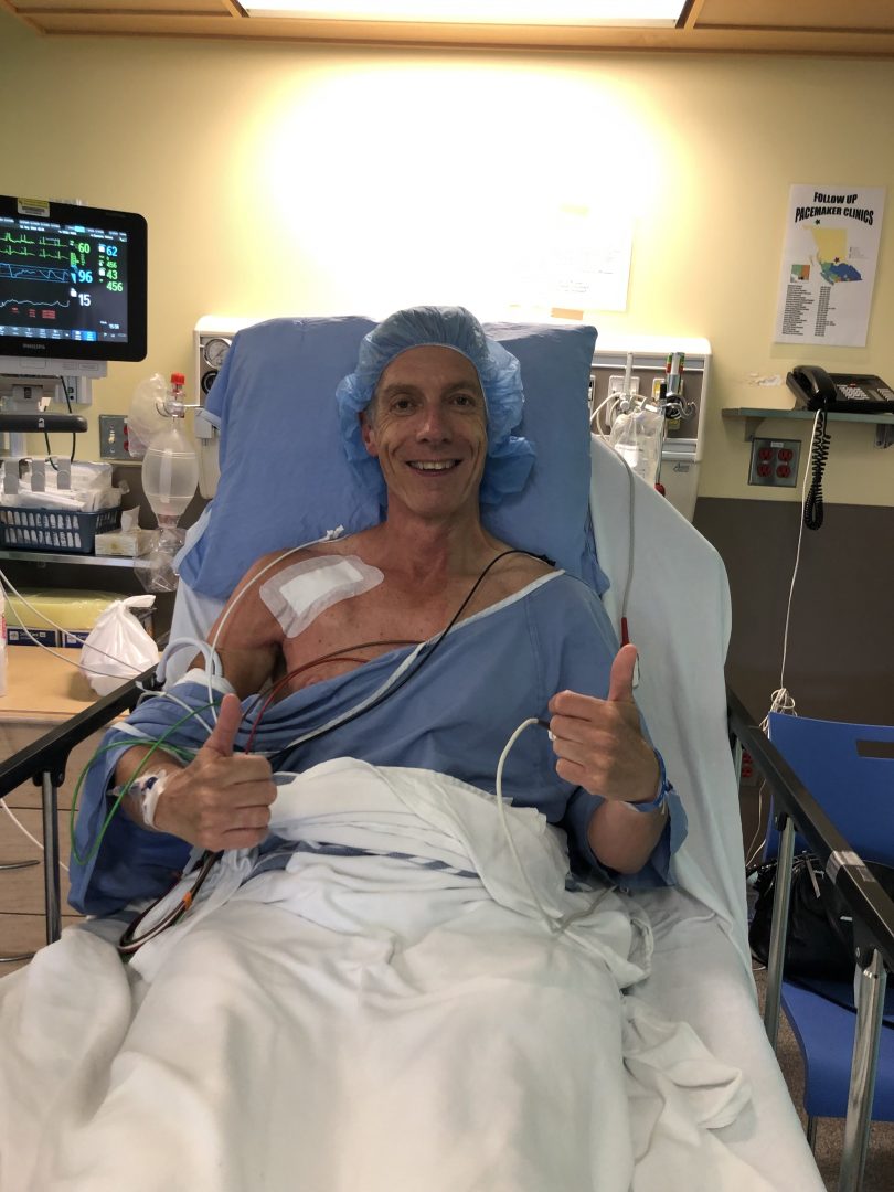 Richard Atkins lying in hospital bed with a smile and two thumbs up.