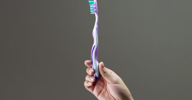 brushing your teeth is an important part of oral care. A hand holds a toothbrush in the center of the photo, there is a plain grey background behind it.
