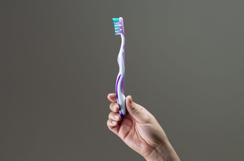 brushing your teeth is an important part of oral care. A hand holds a toothbrush in the center of the photo, there is a plain grey background behind it.